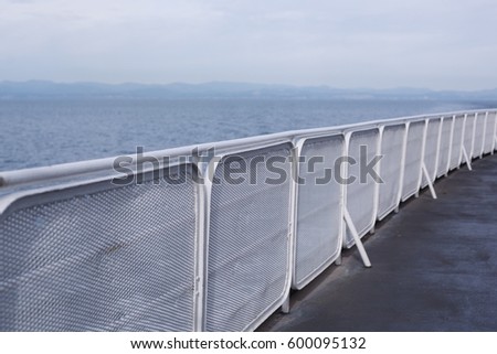 Long oblique perspective of white painted rounded steel pipe railing with curved edge rectangular metal grate on port side of nautical ferry ship at calm blue ocean sea waters under bright hazy sky 