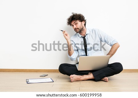 A barefoot businessman sitting on the floor against a wall and using a laptop and showing one side with his pen