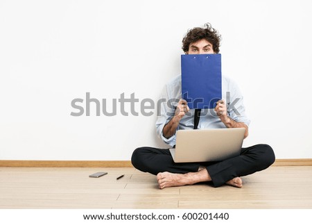 A barefoot businessman sitting on the floor against a wall and using a laptop and covering his face with flip-chart