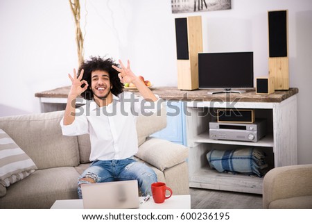 Hipster freelance man showing okay signs to camera while sitting on sofa or couch and working on laptop computer at home alone.
