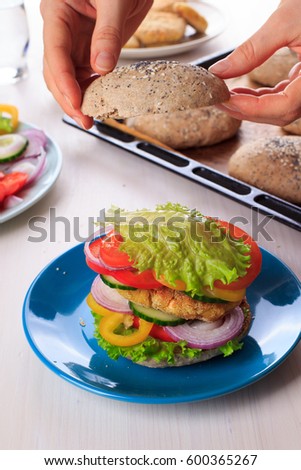 Woman cooking healthy vegan burger with chickpea fritters, vegetables and whole-wheat bun with sesame and poppy seeds on white table for breakfast