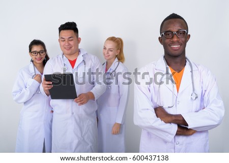 Studio shot of happy young African man doctor with arms crossed and diverse group of multi ethnic doctors smiling while reading on clipboard together