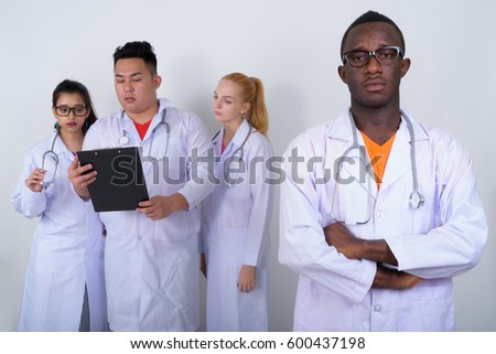 Studio shot of young African man doctor with arms crossed and diverse group of multi ethnic doctors reading on clipboard together