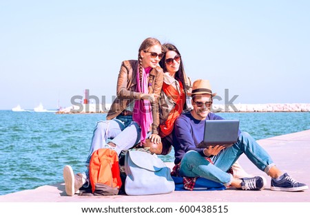 Happy best friends traveling on summer holiday are waiting ferry - Cheerful young travelers  using laptop while sitting by ocean at pier on sunny spring break day - Concept of share journey together