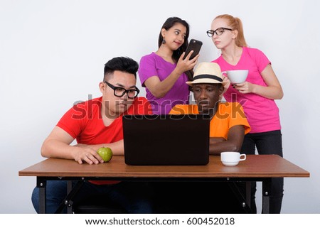 Studio shot of diverse group of multi ethnic friends using laptop with coffee cup and green apple on wooden table together with friends using mobile phone at the back