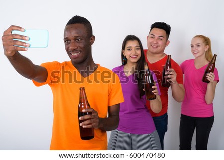 Studio shot of happy young African man with diverse group of multi ethnic friends smiling while holding bottle of beer and taking selfie picture with mobile phone together