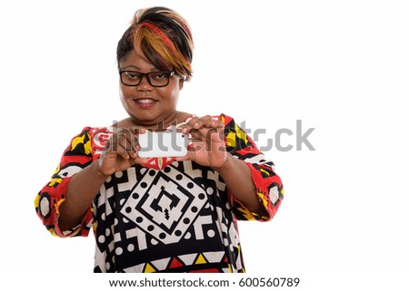 Studio shot of happy fat black African woman smiling while taking picture with mobile phone and looking at camera