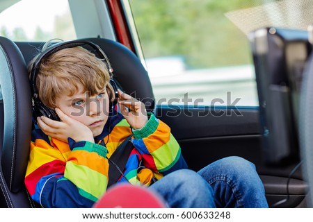 Little blond kid boy watching tv or dvd with headphones during long car driving on family vacations. Leisure for children for long drive. Preschool child sitting in safe car seat.