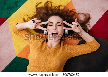 Top view portrait of a young pretty girl in headphones enjoying music while lying on a carpet and showing peace gesture at home with eyes closed