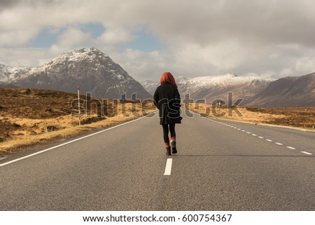 Girl on black clothes wearing a head scarf walks on the middle of a road towards a range of mountains covered in snow in the Glen Coe valley in the Highlands of Scotland, north of Argyll, UK, 