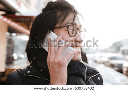 Picture of handsome young asian man wearing glasses and scarf walking outdoors while talking by phone.