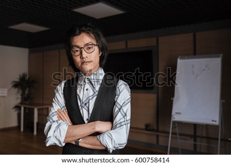 Photo of amazing young asian man wearing glasses dressed in jacket and shirt standing with arms crossed indoors and looking at camera. Coworking concept.