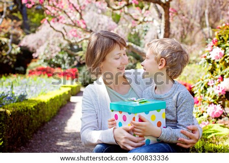 happy young smiling mother holding present from her son in beautiful park with blooming trees at springtime, mother's day holiday concept