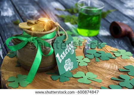 Happy St Patrick's Day concept, Shiny Pot of Gold Leprechaun, clover trefoils, Greeting card with text "Lucky",  tobacco pipe and green ale on wooden stump.