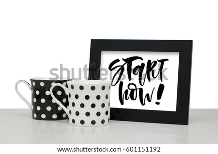 Start now. Handwritten text, inspirational quote. Modern calligraphy. Black wooden frame. Black and white coffee Cup on the table. Close-up, white background