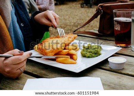 Woman eating fish and chips, traditional English dish, on an pub outdoor wooden table, beer on the background Royalty-Free Stock Photo #601151984