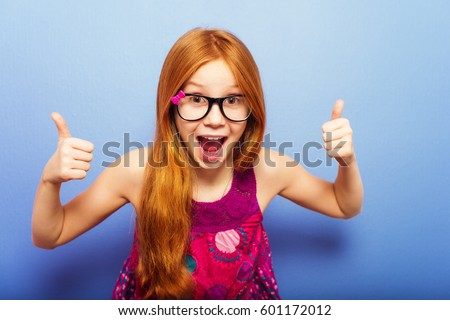 Studio shot of young preteen 9-10 year old redhead girl wearing eyeglasses, standing against blue purple background, big thumbs up, excited facial expression Royalty-Free Stock Photo #601172012