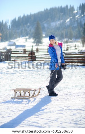Nice young woman pulls sledge. Ground is covered with snow. Serene winter day. Woman smiles. Against the background of a fine winter landscape.