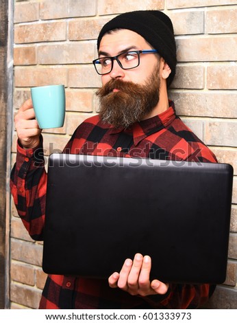 Bearded man, long beard. Brutal caucasian surprised unshaven hipster holding laptop with mag or cup in red black checkered shirt with hat and glasses on beige brick wall studio background