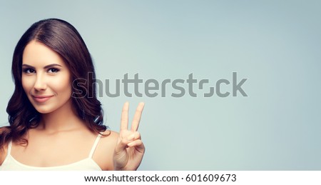 Portrait of beautiful young woman showing two fingers or victory gesture, on grey background. Copyspace area for advertising slogan or text message.