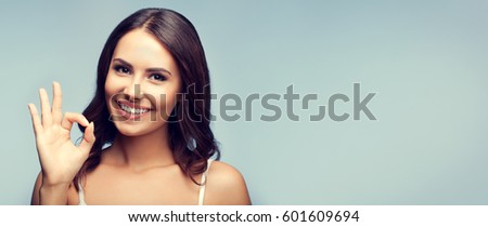 Portrait of young cheerful smiling woman showing okay gesture, over grey background. Copyspace area for advertising slogan or text message.