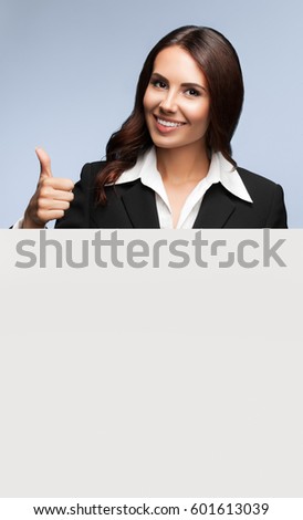 Portrait of happy smiling young businesswoman in black suit, showing blank signboard with blank copyspace area for slogan or text, over grey background, showing thumb up gesture