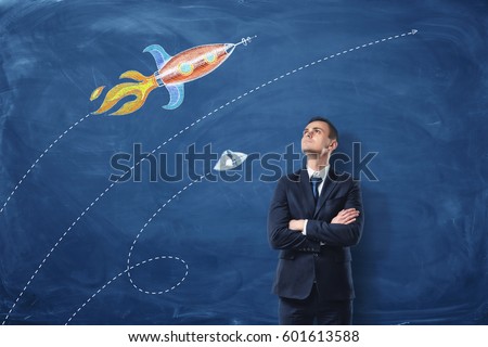 A businessman standing with his arms folded on the dark blue background, he is looking up at the pictures of the rocket and the UFO drawn on the wall. Long-range plans. Flights of imagination. Looking