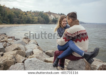 Happy thoughtful couple sitting on a rock beach near sea hugging each other in cold foggy cloudy autumn weather. Copy space