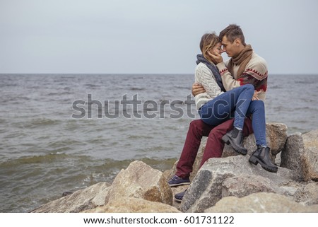 Happy thoughtful couple sitting on a rock beach near sea hugging each other in cold foggy cloudy autumn weather. Copy space