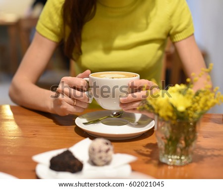 A photo of a coffee cup on a wooden table and a pair of woman's hands holding another cup at the background 