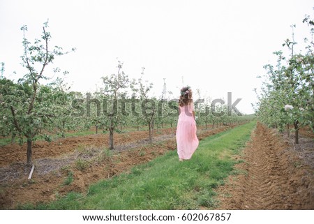 A young girl with a wreath of flowers on her head in a pink chiffon flying dress runs through a flowering garden in the spring. Blossoming pear and apple garden, trees planted in rows