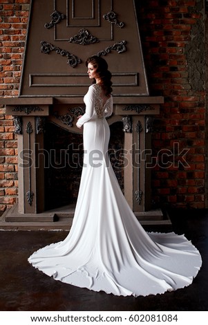 Portrait of a bride, caucasian, brunette girl in white wedding dress with long arm and a cut on her leg, stands in the studio with a stone fireplace with stucco and brick background