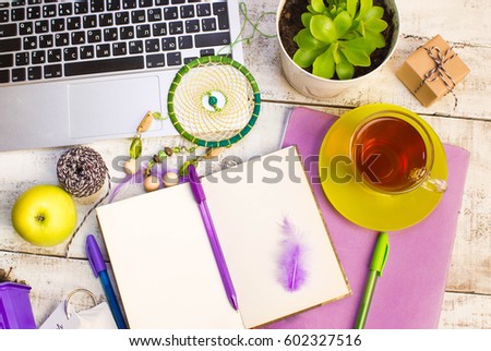 White wooden table with a laptop, a notebook, a mug of tea, pens, flowers, apples.