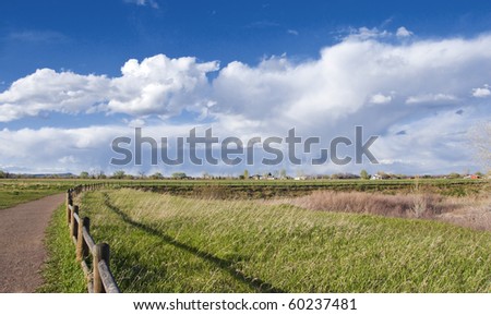 Curved path and rustic railing leads from left to right into the distance of the picture, on the Colorado prairie with dramatic clouds in the background.