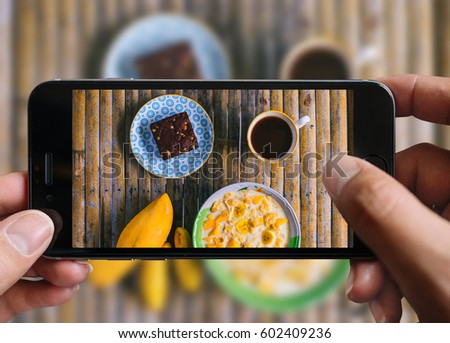 Healthy breakfast concept. Taking picture of cornflakes with milk, banana and mango, coffee cup, brownie with mobile phone. Phone in male hands. Top view, vintage style. Wooden bamboo background.
