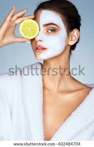 Brunette woman holding a slice of lemon in front of her face. Photo of woman with moisturizing facial mask. Beauty & Skin care concept