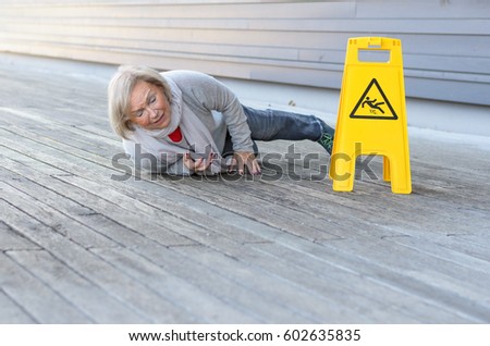 Senior lady slipping and falling on a wet surface with a grimace of pain right alongside a yellow warning sign in German, with foreground copy space