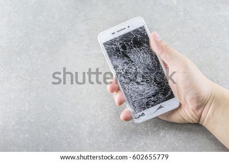 Man's hand holds mobile phone with broken touchscreen on gray background.