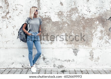 Hipster girl wearing blank gray t-shirt, jeans and backpack posing against rough street wall, minimalist urban clothing style, mockup for tshirt print store Royalty-Free Stock Photo #602669993
