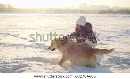 Young beautiful happy girl plays with a retriever dog in the snow in winter in sunny day during sunset time