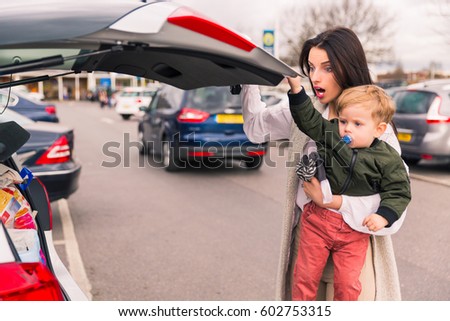 young woman  with a child in her arms, closing the luggage in the car, standing near gray car. After shopping luggage with products