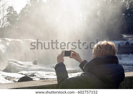 Back view of female tourist photographing frozen waterfall on mobile phone while enjoying her winter holidays. Winter landscape and snow landscapes, frozen waterwall and icicles