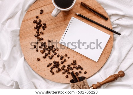 Coffee beans, cup of coffee, cinnamon, notepad with a pencil and coffee pot (turka) on a round wooden board and white cloth. abstract background. Concept photo. View from above
