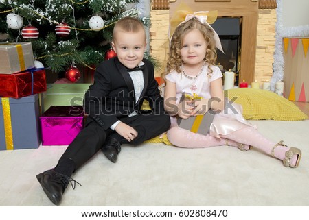 Well-dressed boy and girl sitting near a Christmas tree surrounded by gifts.