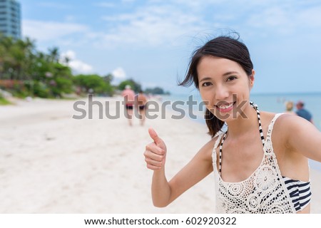 Woman holding camera to take selfie and showing thumb up in sand beach