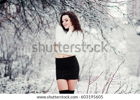 Curly brunette girl background falling snow, wear on black mini skirt and wool stockings. Model on winter. Fashion portrait at snowy weather. toned photo.