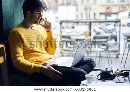 Thoughtful young handsome male photographer talking on phone with girlfriend while sitting in coffee shop using free wireless internet  connection to working with photo editor application on laptop
