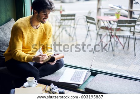 Cheerful male international student in casual outfit sitting in coffee shop preparing homework task making exercises on training website via laptop computer connected to free wireless internet