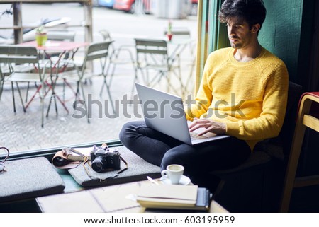 Pensive handsome male blogger updating his profile in social networks with retro photos sharing with followers multimedia files sitting in coffee shop with equipment and laptop computer with wifi