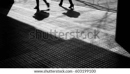 Blurry silhouettes and shadows of two people walking on a city sidewalk, from waist down, in black and white